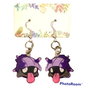 May include: A pair of purple and gray cartoon character earrings with a pink tongue. The earrings are on a white card with floral designs.