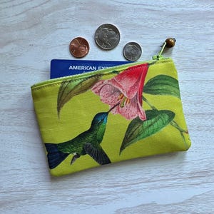 May include: A small, lime green zippered pouch with a hummingbird and flower design. The hummingbird is green and blue, and the flower is pink and red. The pouch is open, revealing a blue credit card and several coins.