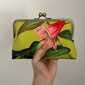 May include: A lime green clutch with a hummingbird and flower print. The clutch has a metal clasp and a floral design featuring a pink flower and a blue and green hummingbird. The clutch is held in a hand.