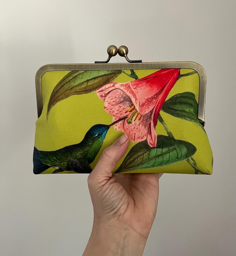 May include: A lime green clutch with a hummingbird and flower print. The clutch has a metal clasp and a floral design featuring a pink flower and a blue and green hummingbird. The clutch is held in a hand.