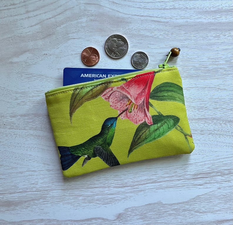 May include: A small, lime green zippered pouch with a hummingbird and flower design. The hummingbird is green and blue, and the flower is pink and red. The pouch is open, revealing a blue credit card and several coins.