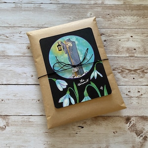 May include: A package wrapped in brown paper, tied with black twine. A black card with a tarot illustration of The Hermit, a robed figure with a lantern, is affixed to the front. The card also features snowdrop flowers and the number 9.