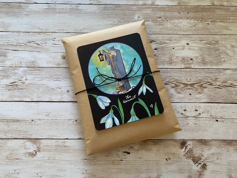 May include: A package wrapped in brown paper, tied with black twine. A black card with a tarot illustration of The Hermit, a robed figure with a lantern, is affixed to the front. The card also features snowdrop flowers and the number 9.