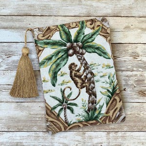 May include: A rectangular book cover featuring a tropical scene with a monkey climbing a palm tree, with green leaves and brown coconuts. The cover has a beige tassel attached to the top.