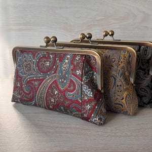 May include: Three paisley print clutch purses with bronze-colored metal frames. The purses feature a variety of colors, including red, blue, gold, and black. The purses are arranged in a row, with the first purse in focus.