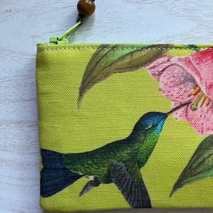 May include: A lime green zippered pouch featuring a vibrant print of a hummingbird and pink flowers. The hummingbird is depicted in shades of green and blue, with a long beak. The zipper pull has a wooden bead.
