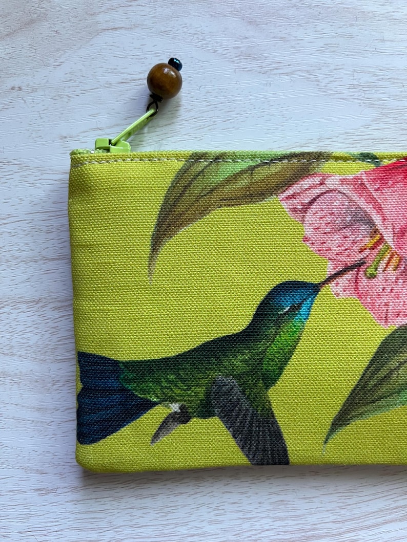 May include: A lime green zippered pouch featuring a vibrant print of a hummingbird and pink flowers. The hummingbird is depicted in shades of green and blue, with a long beak. The zipper pull has a wooden bead.