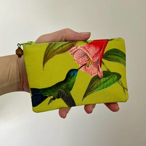 May include: A small, rectangular pouch with a vibrant yellow background. The pouch features a detailed illustration of a green and blue hummingbird feeding from a pink flower. A wooden bead adorns the zipper pull.