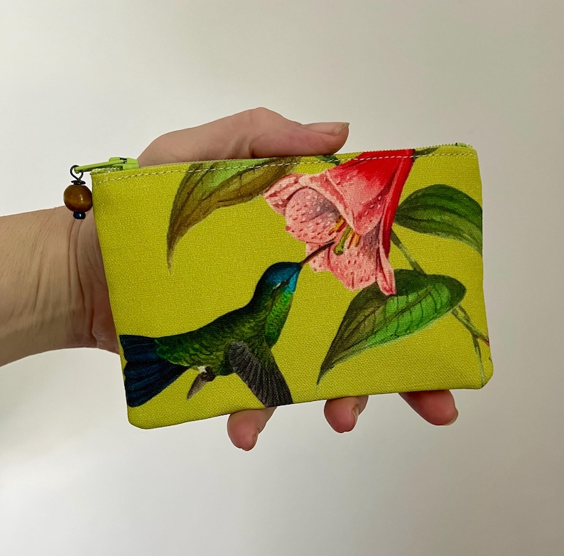May include: A small, rectangular pouch with a vibrant yellow background. The pouch features a detailed illustration of a green and blue hummingbird feeding from a pink flower. A wooden bead adorns the zipper pull.