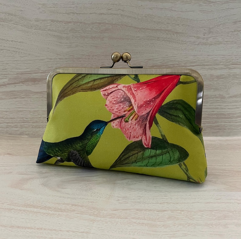 May include: A rectangular clutch with a metal clasp. The clutch features a vibrant print of a hummingbird, a pink flower, and green leaves on a lime green background. The hummingbird has blue and green feathers.
