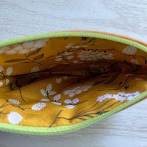 May include: Open view of a small, zippered pouch. The exterior is orange, with a bright green zipper. The interior lining is a mustard yellow fabric with a white floral pattern and black stems.