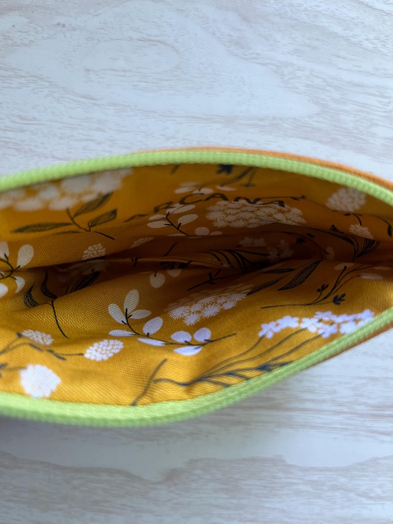 May include: Open view of a small, zippered pouch. The exterior is orange, with a bright green zipper. The interior lining is a mustard yellow fabric with a white floral pattern and black stems.