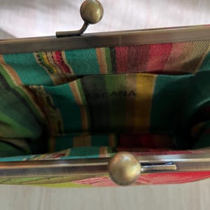 May include: A close-up of a vintage-style purse with a metal clasp and a colorful striped interior. The purse has a brass-colored frame and clasp. The interior fabric features green, red, yellow, and white stripes. The word "ARCANA" is visible inside.