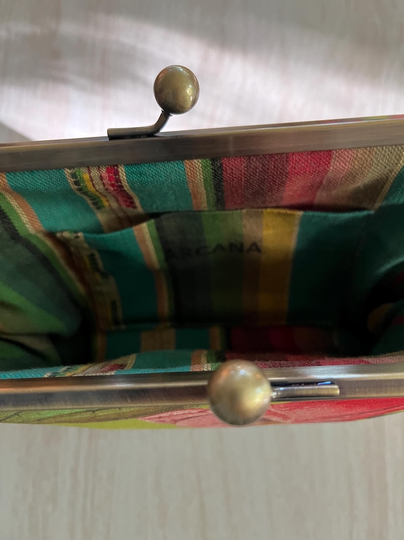 May include: A close-up of a vintage-style purse with a metal clasp and a colorful striped interior. The purse has a brass-colored frame and clasp. The interior fabric features green, red, yellow, and white stripes. The word "ARCANA" is visible inside.