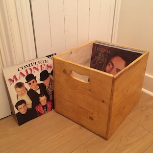 May include: A light-stained wooden crate, designed to hold vinyl records, with a handle cut-out. A record sleeve with the text "COMPLETE MADNESS" leans against the crate. Inside, a "LEGEND BOB MARLEY" album is visible. The crate sits on a wooden floor.