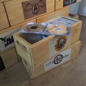 May include: Wooden storage crates with band logos, including Aerosmith, Guns N' Roses, and Whitesnake. Several CDs are visible inside one crate. The crates are stacked, and the wood has a natural, light colour.