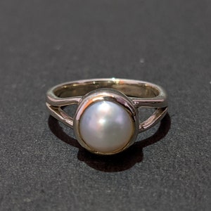 Puede incluir: Un anillo de plata con una perla blanca engastada en un engaste de bisel. El anillo tiene un diseño simple y elegante.