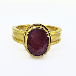 May include: A gold ring with a large, oval-shaped ruby gemstone set in a bezel setting. The ring band has a textured, grooved design.
