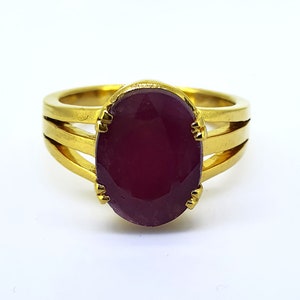 May include: A gold ring featuring a large, oval-shaped ruby gemstone set in the centre. The ring band has three thin bands of gold.
