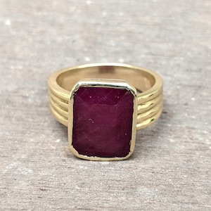 Peut inclure: Une bague en or avec une grande pierre précieuse rectangulaire rouge foncé sertie au centre. La bande de la bague est épaisse et présente un motif texturé.