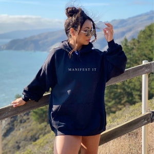 Op de afbeelding: Een zwarte oversized hoodie met de tekst "MANIFEST IT" in wit gedrukt op de voorkant. De hoodie wordt gedragen door een persoon die buiten staat tegen een achtergrond van een blauwe lucht, oceaan en groene heuvels.