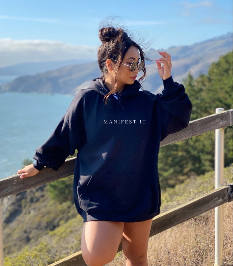 Op de afbeelding: Een zwarte oversized hoodie met de tekst "MANIFEST IT" in wit gedrukt op de voorkant. De hoodie wordt gedragen door een persoon die buiten staat tegen een achtergrond van een blauwe lucht, oceaan en groene heuvels.