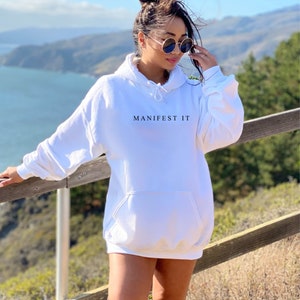 Op de afbeelding: Een witte oversized hoodie met de tekst "MANIFEST IT" bedrukt op de voorkant. De hoodie heeft een capuchon met trekkoord en een kangoeroezak.