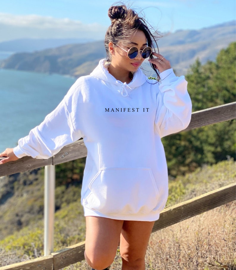 Op de afbeelding: Een witte oversized hoodie met de tekst "MANIFEST IT" bedrukt op de voorkant. De hoodie heeft een capuchon met trekkoord en een kangoeroezak.