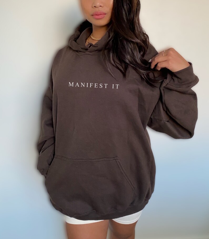 Op de afbeelding: Een oversized bruine hoodie met een witte geborduurde tekst "MANIFEST IT" op de voorkant.