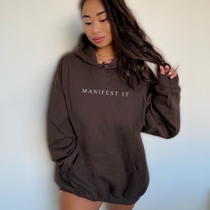 Op de afbeelding: Een oversized bruine hoodie met de tekst "MANIFEST IT" in witte letters op de voorkant.