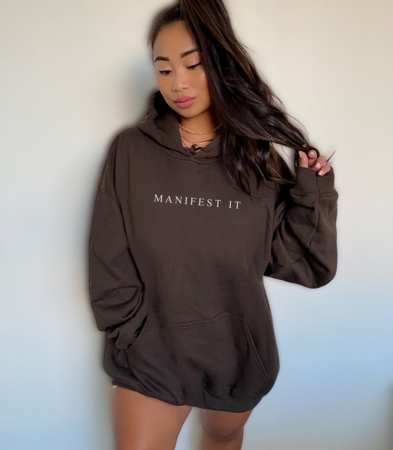 Op de afbeelding: Een oversized bruine hoodie met de tekst "MANIFEST IT" in witte letters op de voorkant.