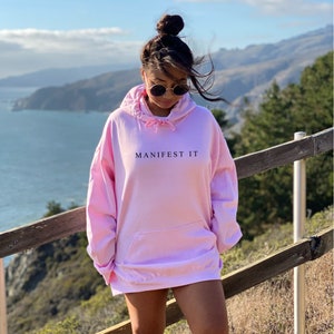 Op de afbeelding: Een roze oversized hoodie met de tekst "MANIFEST IT" erop gedrukt. De hoodie wordt gedragen door iemand die op een houten railing staat met uitzicht op een waterpartij.