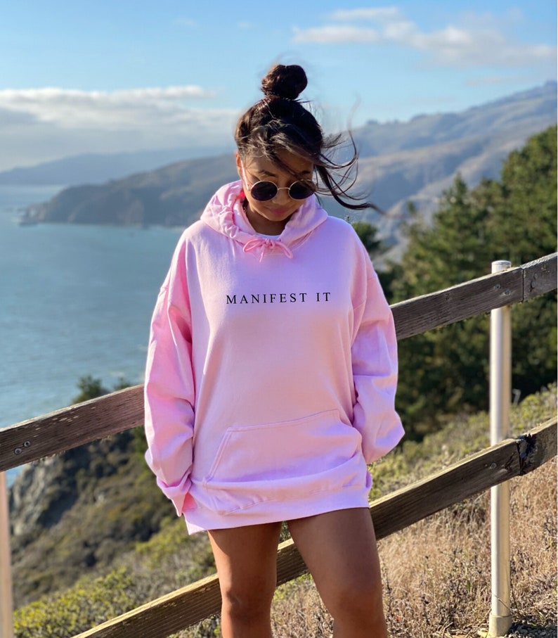 Op de afbeelding: Een roze oversized hoodie met de tekst "MANIFEST IT" erop gedrukt. De hoodie wordt gedragen door iemand die op een houten railing staat met uitzicht op een waterpartij.