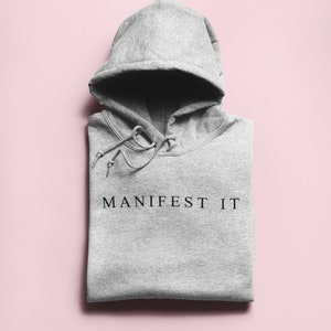 Op de afbeelding: Grijze hoodie met de tekst "MANIFEST IT" in het zwart op de voorkant.