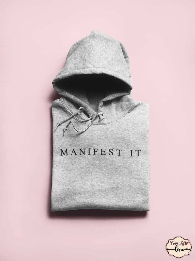 Op de afbeelding: Grijze hoodie met de tekst "MANIFEST IT" in het zwart op de voorkant.