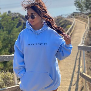 Op de afbeelding: Een lichtblauw hoodie met de tekst "MANIFEST IT" in het zwart op de voorkant.