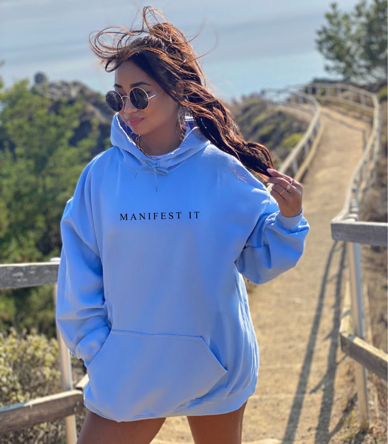Op de afbeelding: Een lichtblauw hoodie met de tekst "MANIFEST IT" in het zwart op de voorkant.
