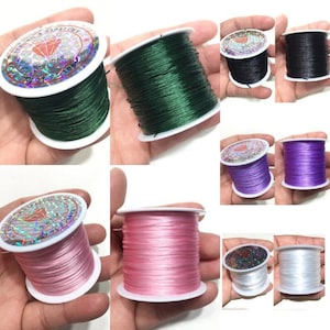 May include: Spools of colorful, stretchy string in various colors, including green, black, purple, pink, and white. Each spool is on a white plastic base with a holographic label. The string is tightly wound, suggesting it's for jewelry making or crafts.