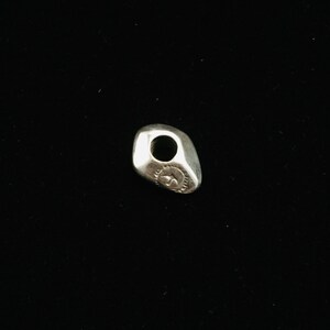 Op de afbeelding: Een zilveren bedel in de vorm van een gladde, ronde steen met een gat in het midden. De bedel heeft een klein, gegraveerd ontwerp aan &eacute;&eacute;n kant.