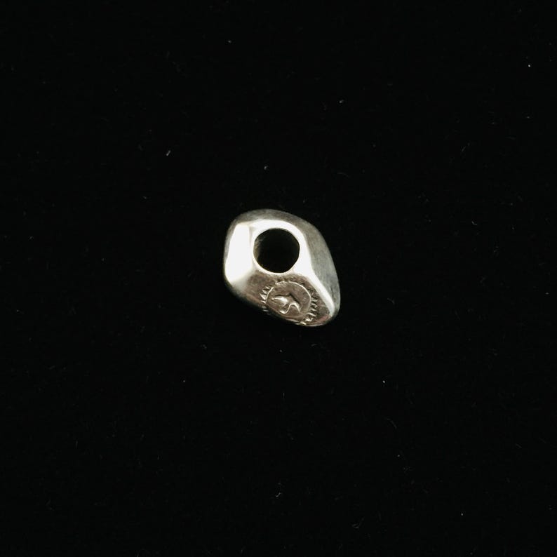 Op de afbeelding: Een zilveren bedel in de vorm van een gladde, ronde steen met een gat in het midden. De bedel heeft een klein, gegraveerd ontwerp aan &eacute;&eacute;n kant.