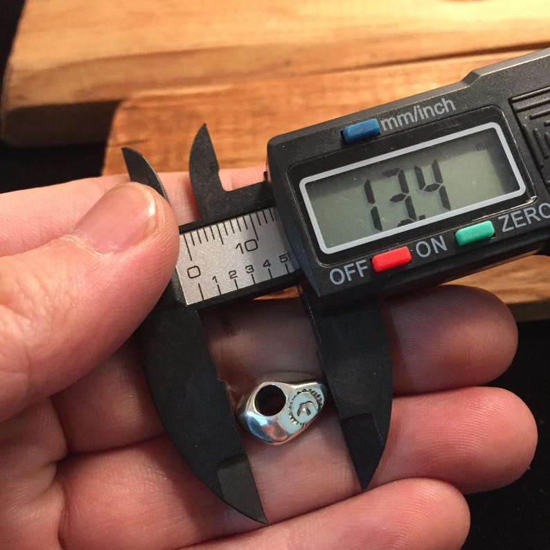Op de afbeelding: Een zilveren schedelbedel met een klein gaatje bovenaan, gemeten met een digitale schuifmaat die 1,34 inch aangeeft.