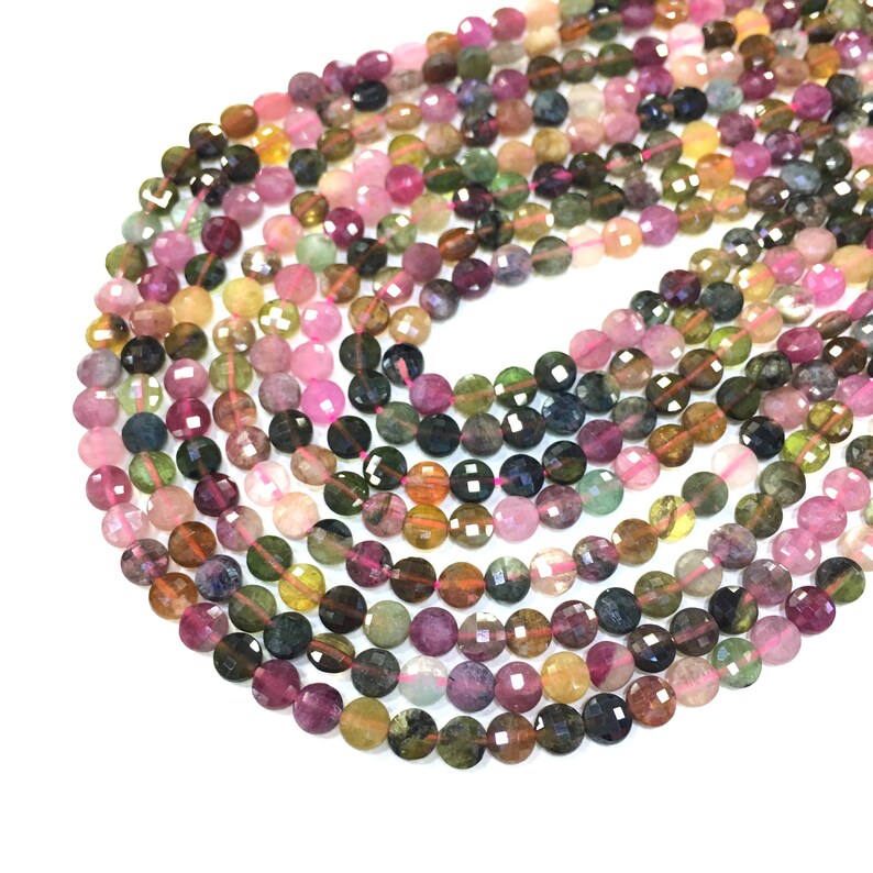 May include: A strand of multi-colored round gemstone beads. The beads are faceted and have a variety of colors, including pink, green, yellow, and blue. The beads are strung on a thin, pink thread.