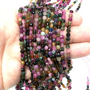 May include: A strand of multi-colored faceted round gemstone beads. The beads are a mix of pink, green, black, and brown. The beads are strung on a clear elastic thread.