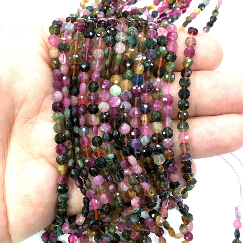 May include: A strand of multi-colored faceted round gemstone beads. The beads are a mix of pink, green, black, and brown. The beads are strung on a clear elastic thread.