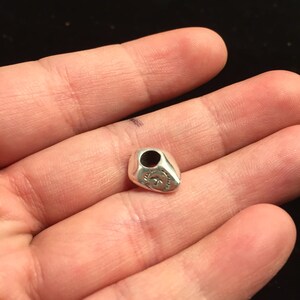 Op de afbeelding: Een zilveren bedel in de vorm van een traan met een gat aan de bovenkant om aan een armband of ketting te bevestigen. De bedel heeft een ontwerp aan de voorkant dat lijkt op een gestileerde bloem.