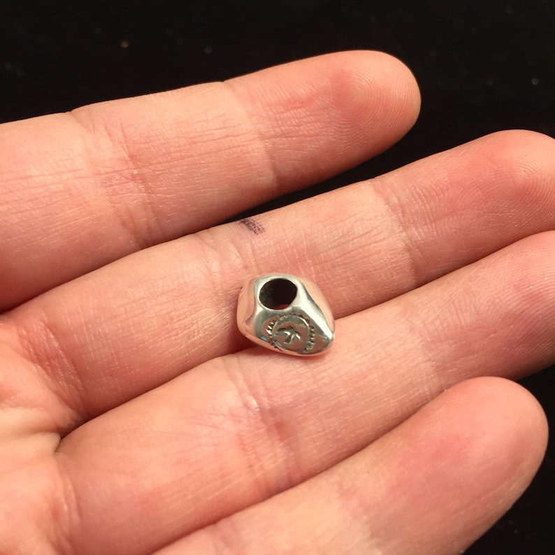 Op de afbeelding: Een zilveren bedel in de vorm van een traan met een gat aan de bovenkant om aan een armband of ketting te bevestigen. De bedel heeft een ontwerp aan de voorkant dat lijkt op een gestileerde bloem.