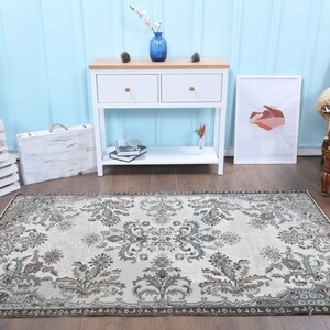 May include: A vintage style rug with a cream background and a floral pattern in shades of brown and green. The rug is laid out on a wooden floor in a room with white furniture and a blue wall.