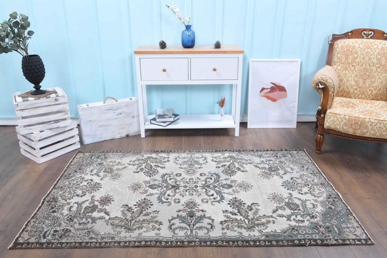 May include: A vintage style rug with a cream background and a floral pattern in shades of brown and green. The rug is laid out on a wooden floor in a room with white furniture and a blue wall.