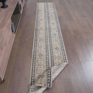 Peut inclure: Un long tapis de couloir étroit présentant une base de couleur crème avec des motifs floraux et cachemire répétitifs complexes dans des tons de bleu, de beige et de brun. Le tapis a un motif de bordure rayé distinct et est posé à plat.