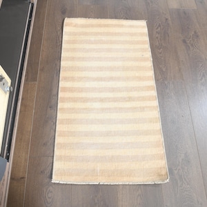 May include: A rectangular rug with horizontal stripes in shades of beige and tan. The rug is placed on a wooden floor. The rug's design is simple and neutral, suitable for various interior styles.